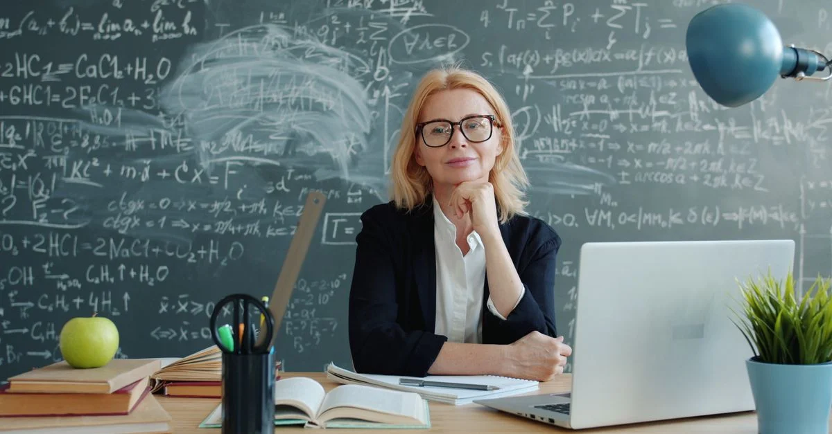 Une enseignante expérimentée est assise devant un tableau noir avec des formules mathématiques.